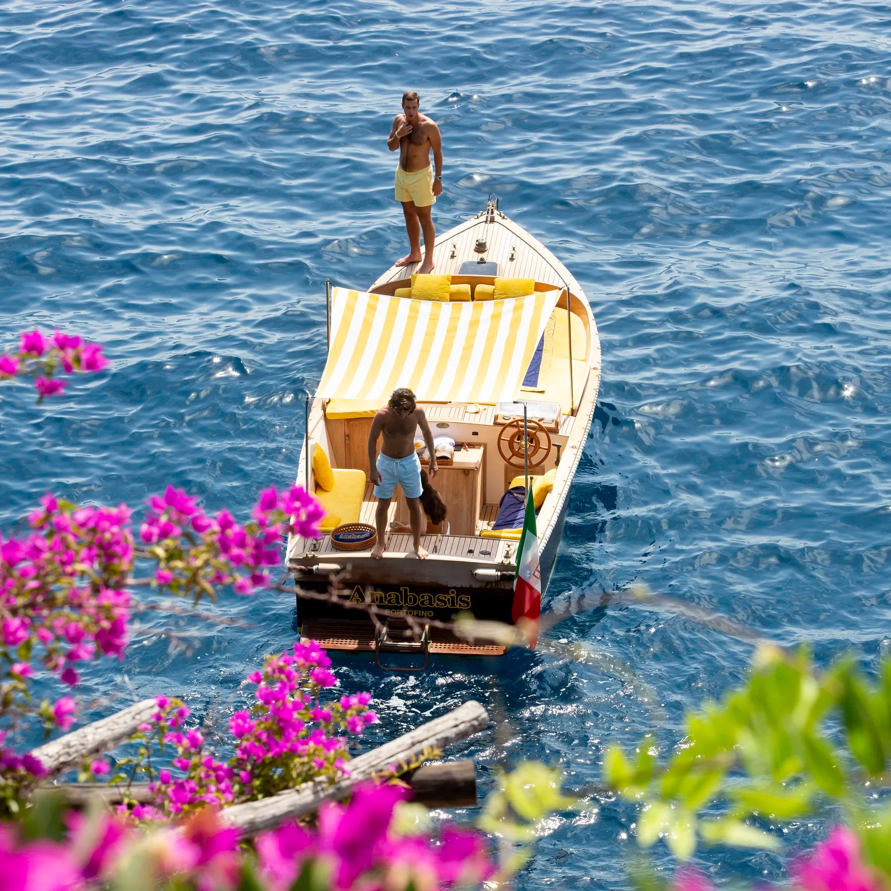 Two people on a boat in the middle of a body of water with flowers and greenery in the foreground.