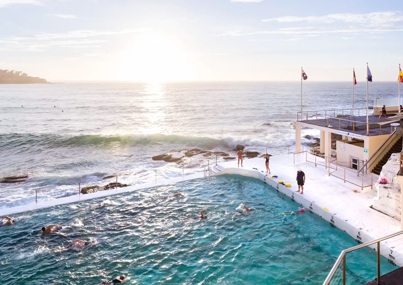 People swimming in an oceanside pool with ocean view and lifeguard tower.