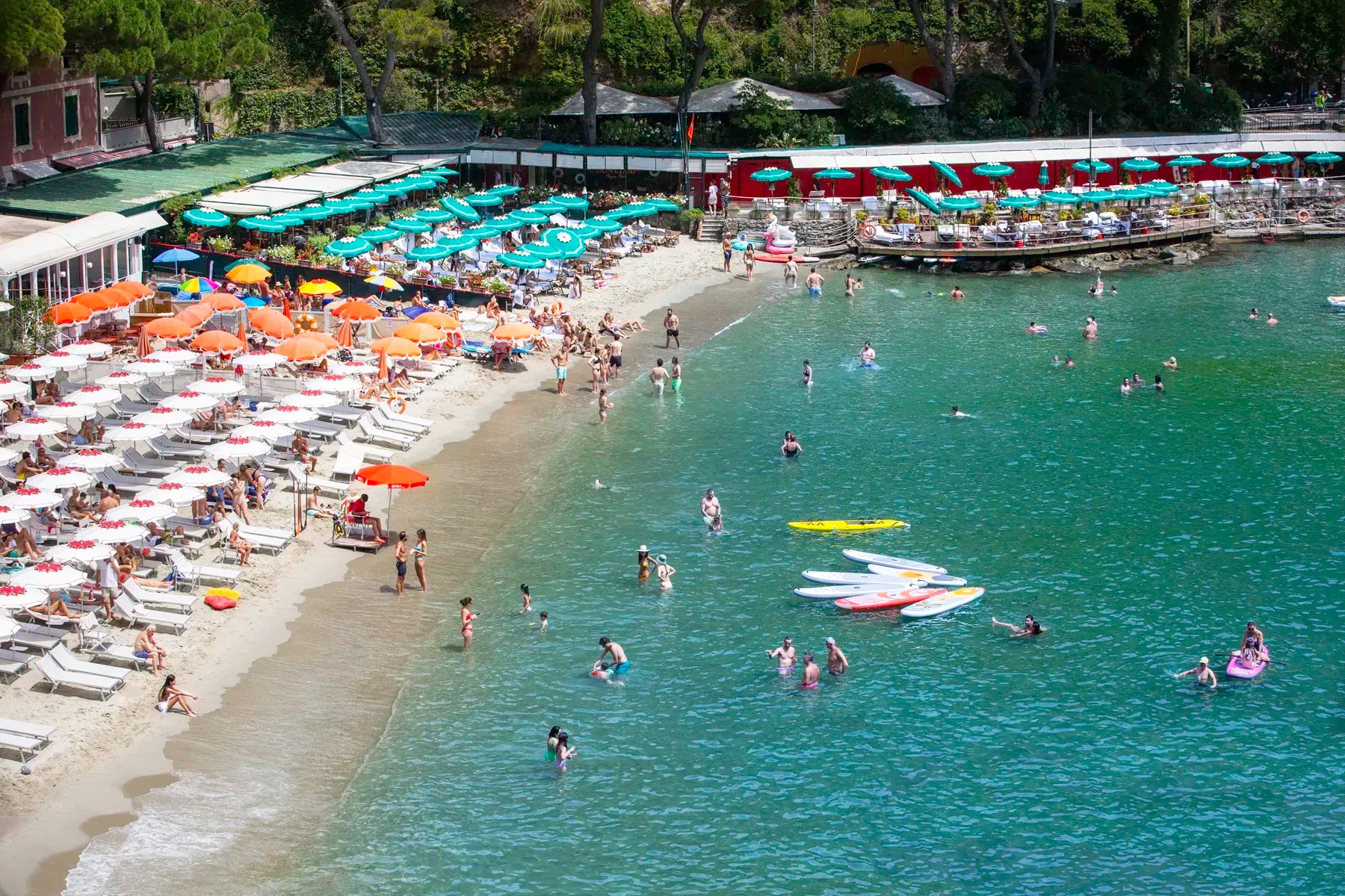 Beach scene with people swimming and sunbathing, surrounded by greenery.