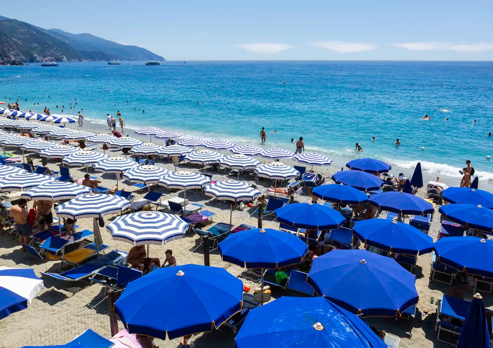 Beach scene with numerous blue and white umbrellas, people, and a clear blue sky.