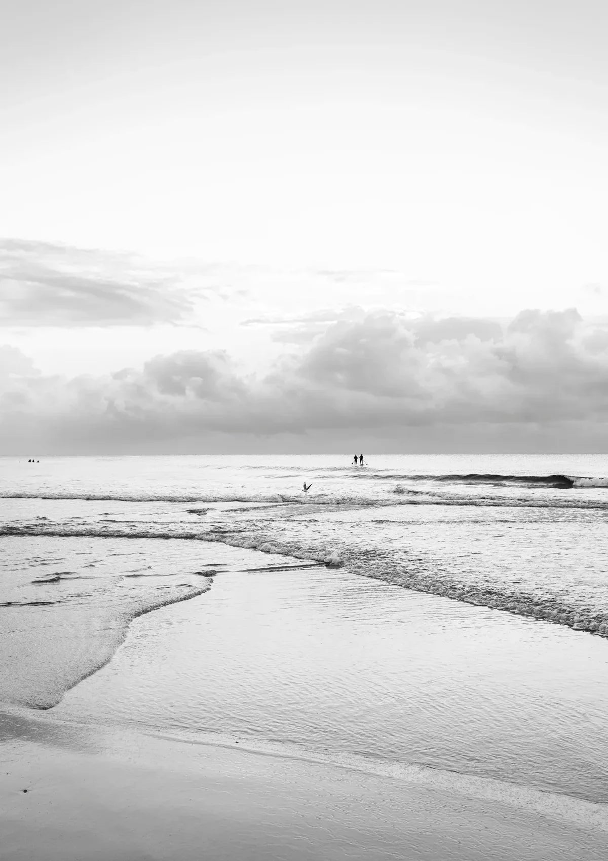Black and white beach scene with people walking on the horizon under a cloudy sky.