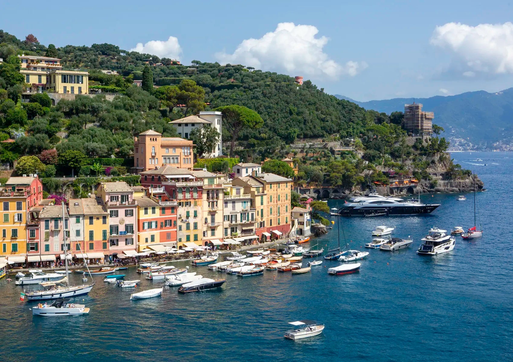 Coastal town with colorful buildings and boats in a harbor, surrounded by green hills and blue sky.