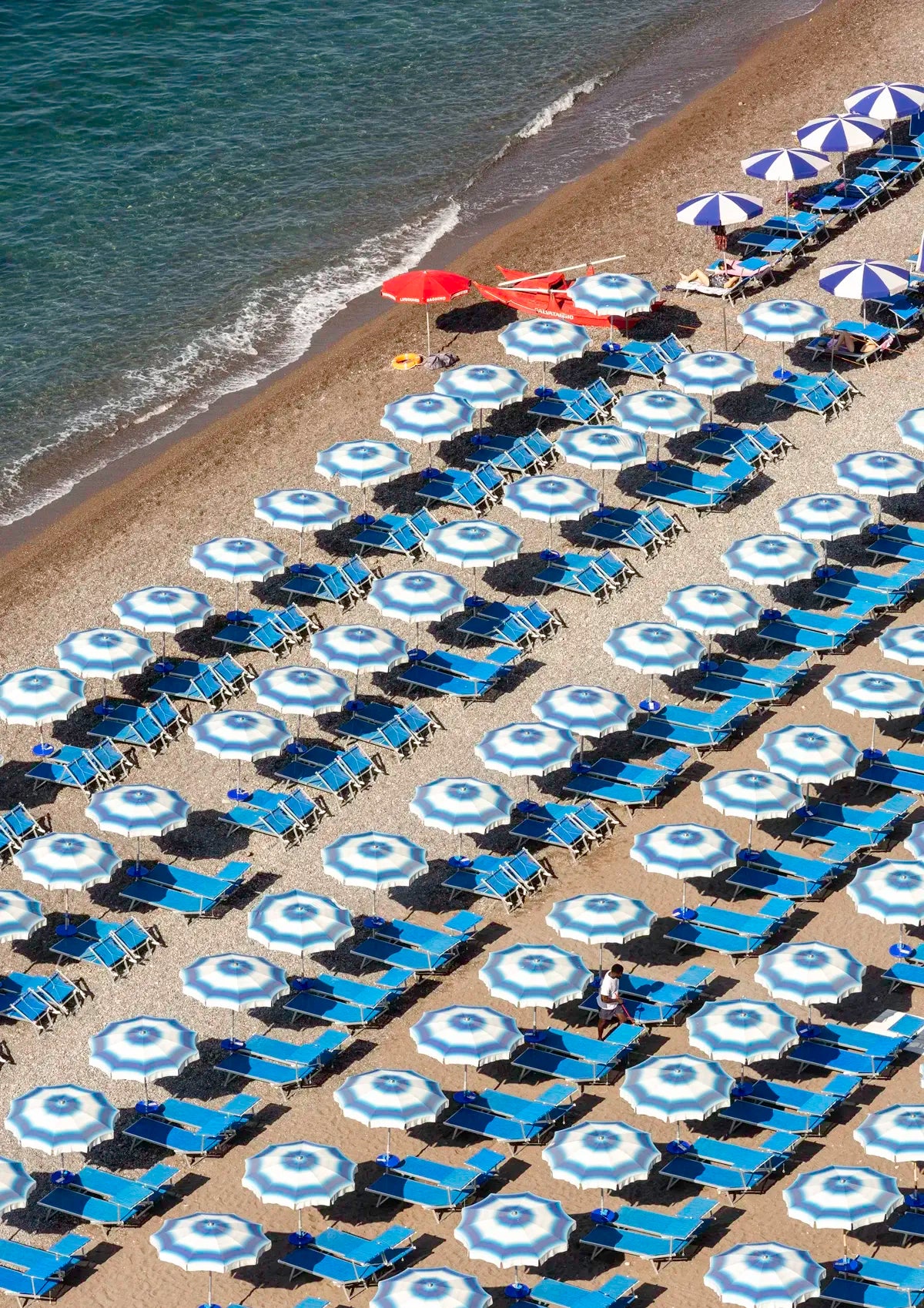 Beach scene with rows of blue and white umbrellas and lounge chairs on a sandy shore.