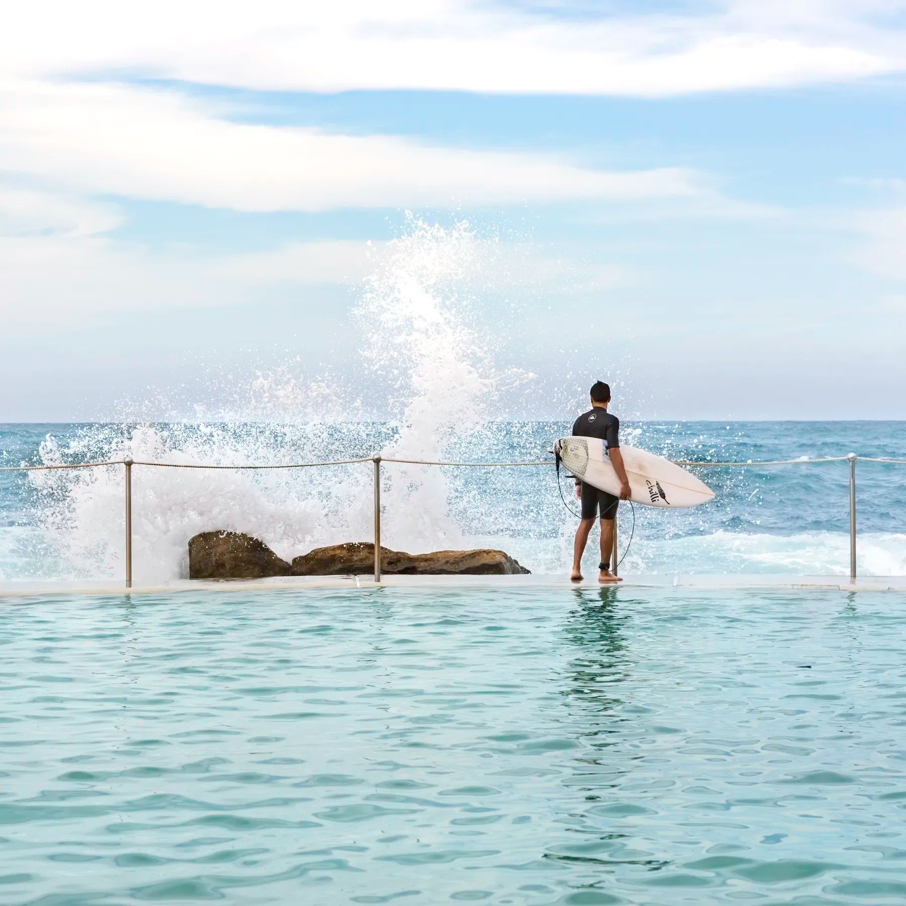 Person holding a surfboard standing in shallow water with a large wave crashing behind them.