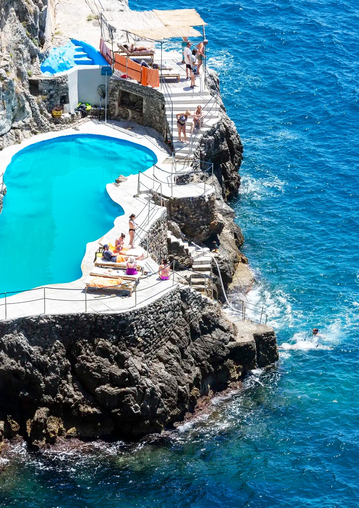 People enjoying a pool built into a rocky cliffside with ocean view.