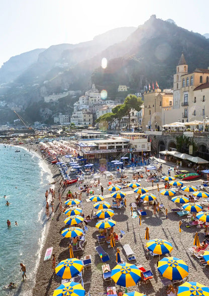 Beach scene with people, umbrellas, and buildings on a mountainous island.
