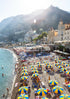 Beach scene with people, umbrellas, and buildings on a mountainous island.