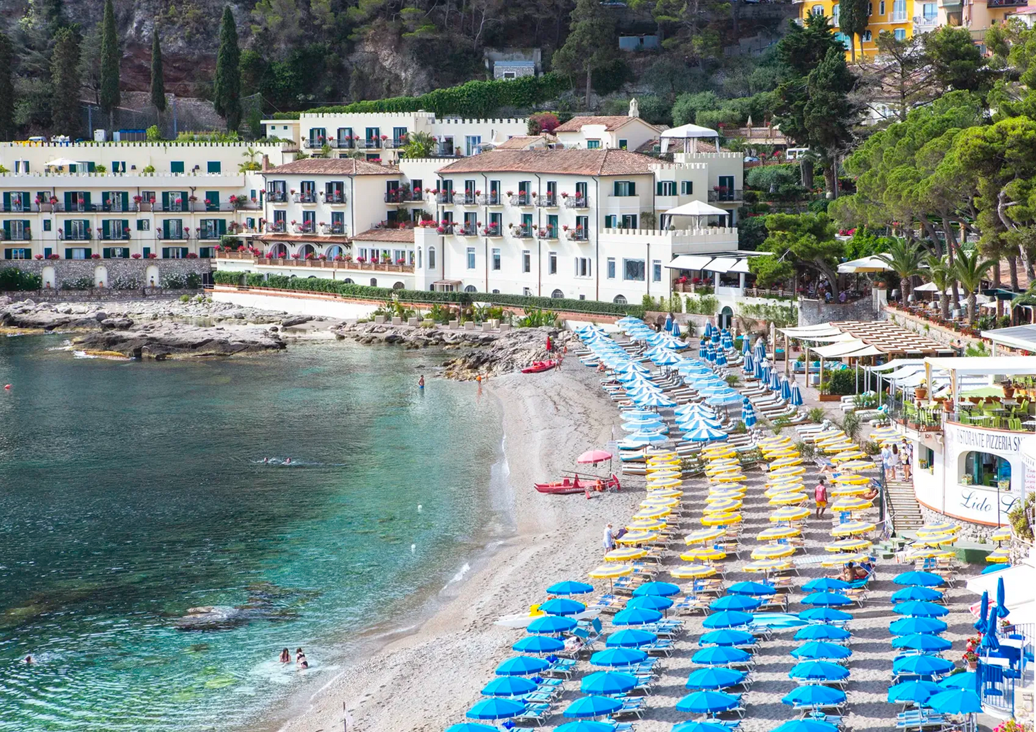 Beach scene with colorful umbrellas, buildings, and trees along a coastal area.
