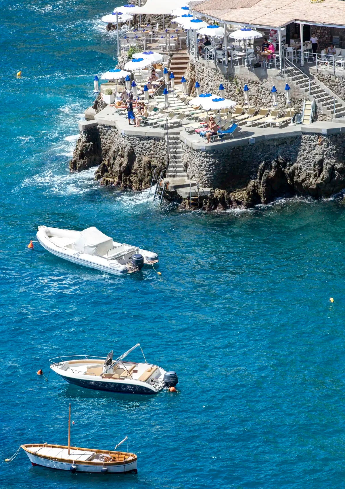 Boats in the water below a cliffside italian beachclub with umbrellas and sunbeds and people relaxing