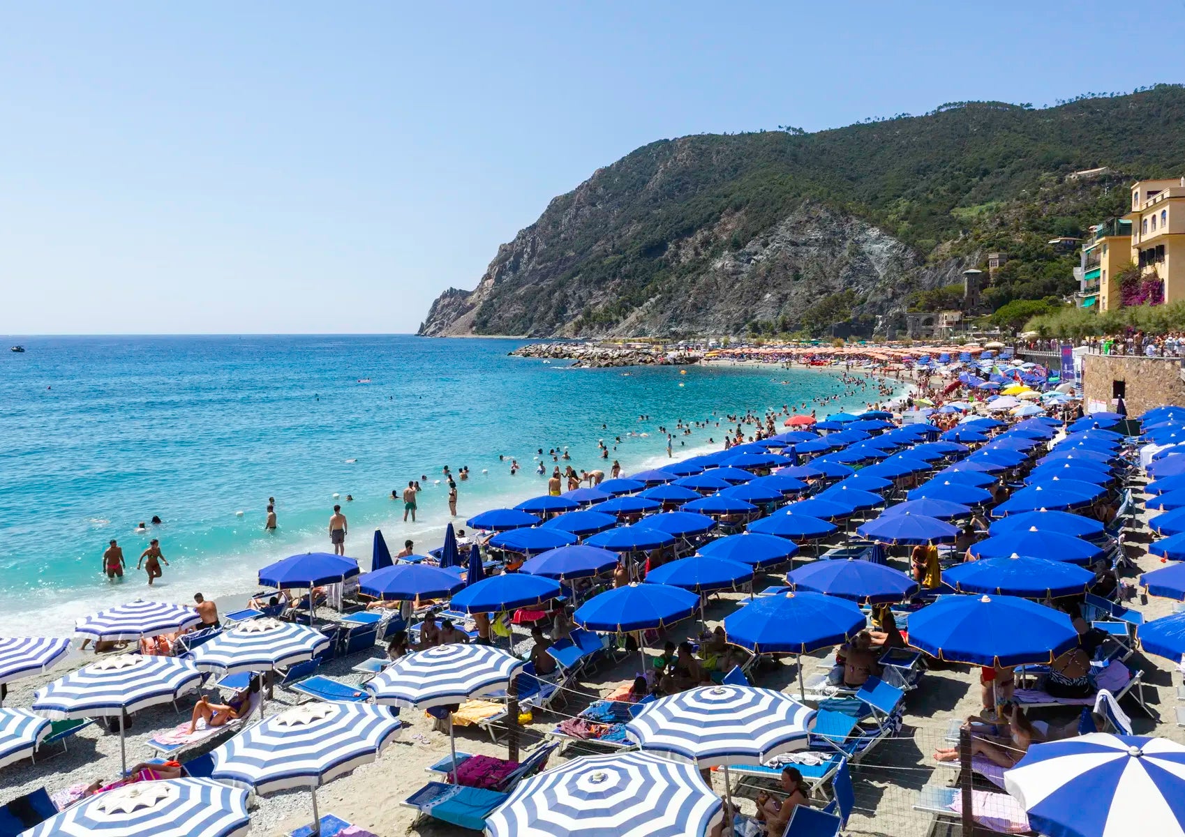 Beach scene with blue and white umbrellas, people, and a mountainous background.