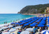 Beach scene with blue and white umbrellas, people, and a mountainous background.