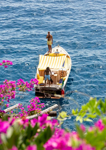 Two people on a small boat in the middle of a body of water with flowers and greenery in the foreground.