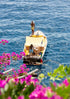 Two people on a small boat in the middle of a body of water with flowers and greenery in the foreground.