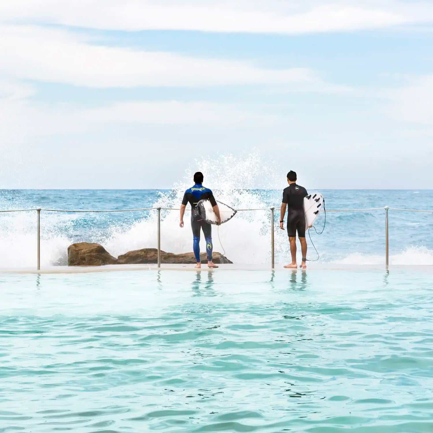 Two surfers with their boards standing in shallow water near rocks.