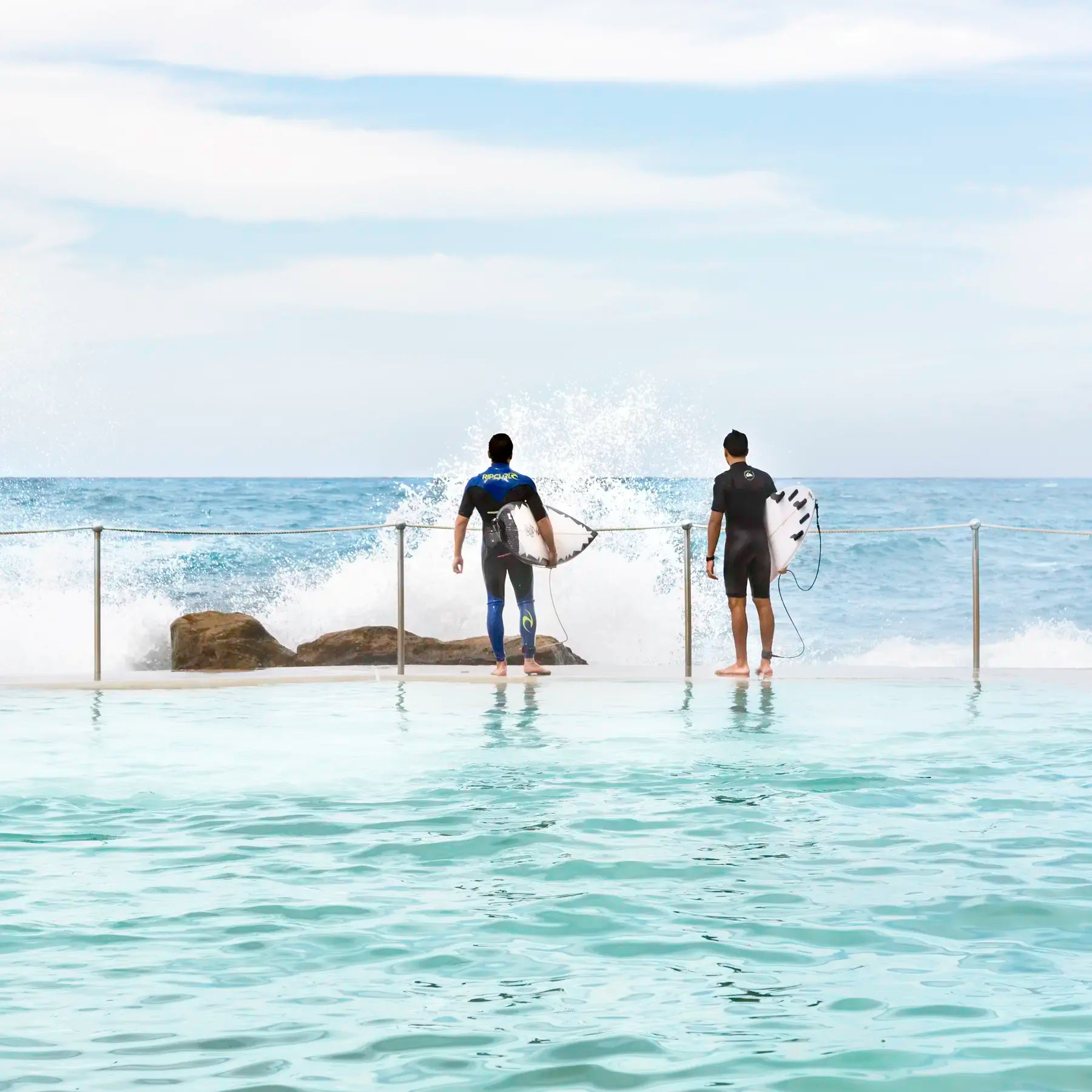 Two surfers with their boards standing in shallow water near rocks.