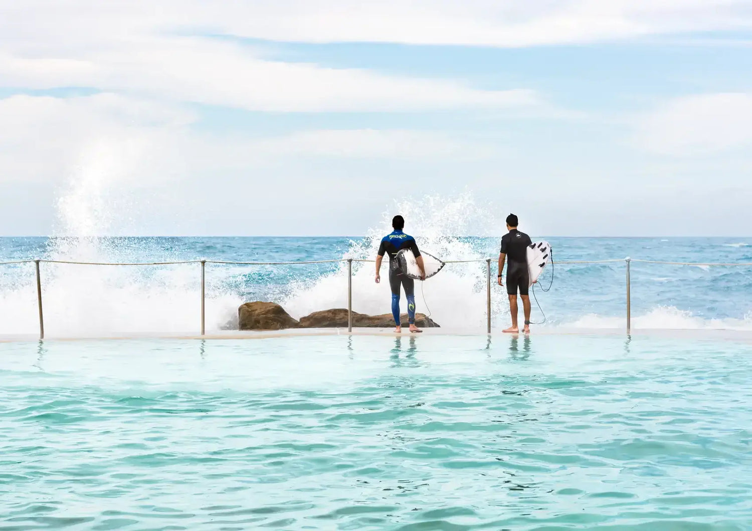 Two people standing in a pool with ocean view, surrounded by water and rocks.