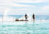 Two people standing in a pool with ocean view, surrounded by water and rocks.