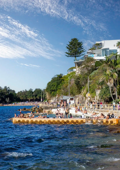 People enjoying a sunny day at a waterfront with modern buildings and greenery.