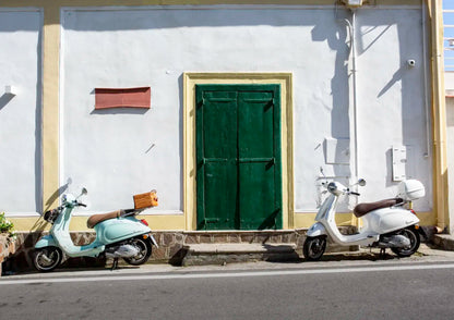 Two scooters parked on a street in front of a white wall with a green door.