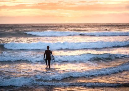Person in a wetsuit walking into the ocean with a surfboard at sunset.
