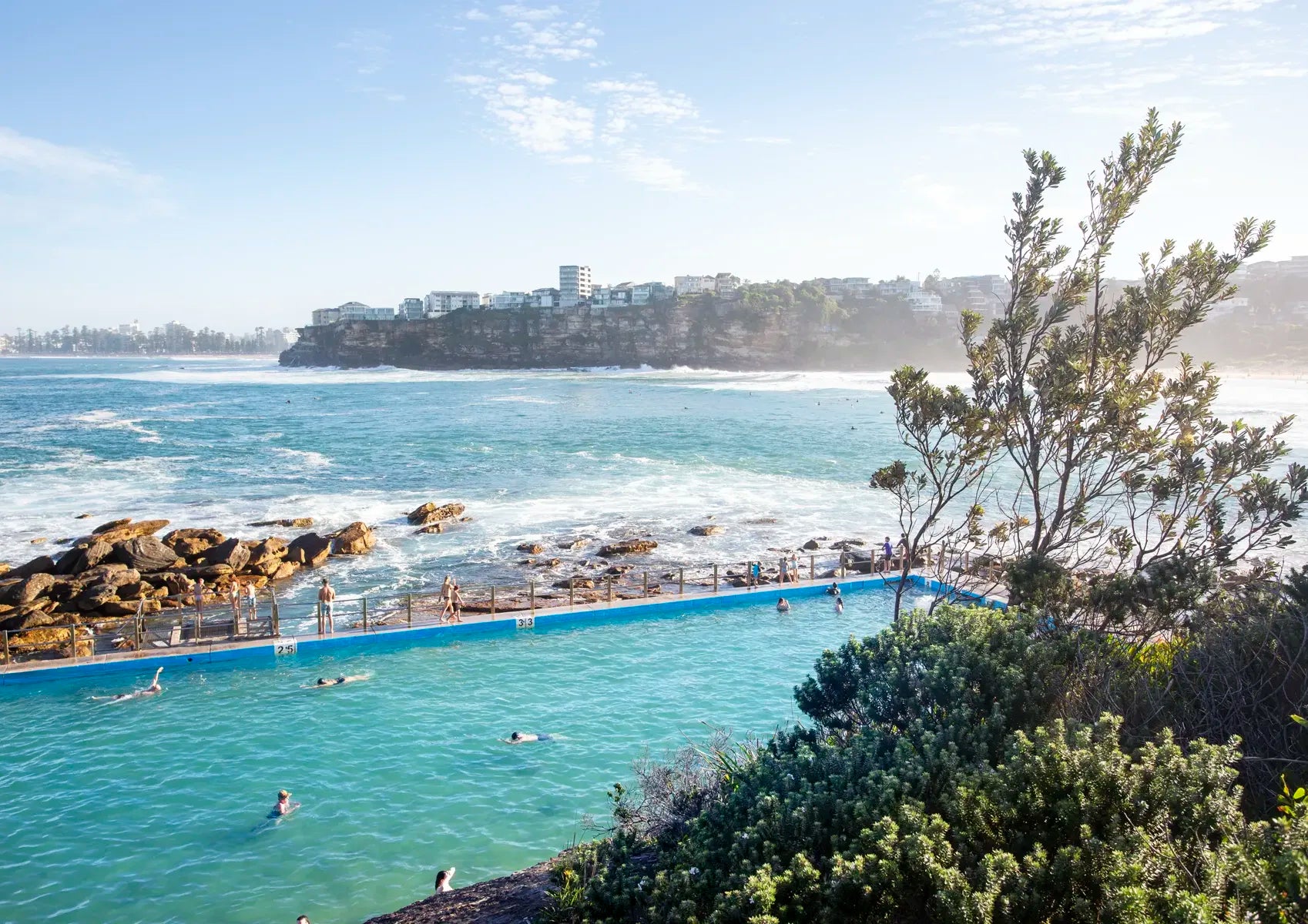 Open-air pool by the ocean with people swimming, surrounded by greenery and a clear sky.