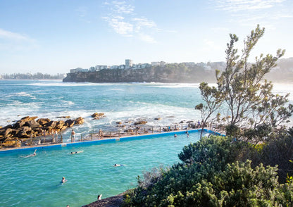 Open-air pool by the ocean with people swimming, surrounded by greenery and a clear sky.