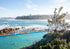 Open-air pool by the ocean with people swimming, surrounded by greenery and a clear sky.