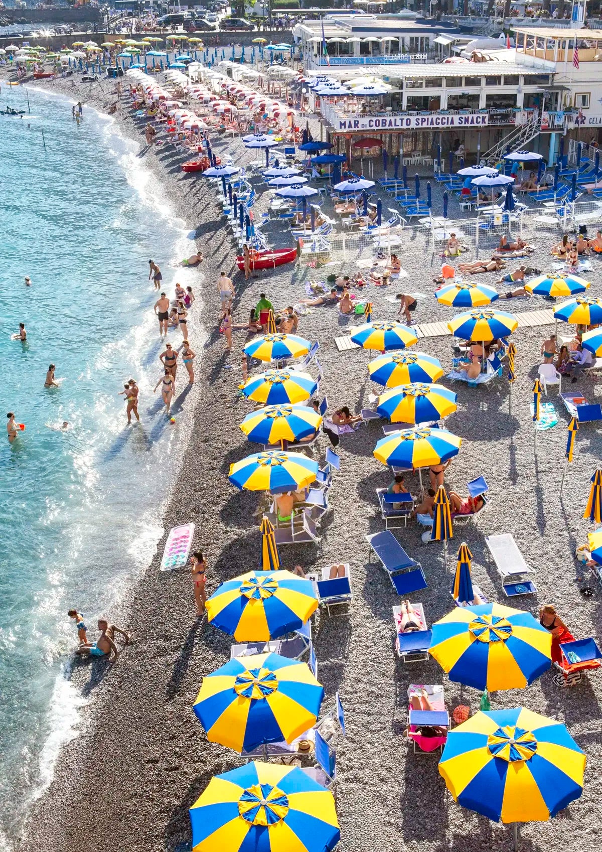 Beach scene with blue and yellow umbrellas, people, and buildings.