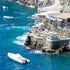Coastal scene at a beach club with a restaurant by the sea, boats in the water, and people enjoying the view from the cliffs.