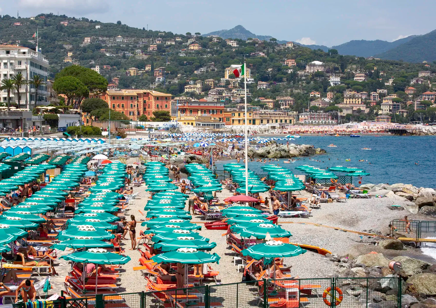 Beach scene with rows of umbrellas and chairs, buildings, and mountains in the background.