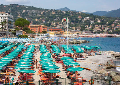 Beach scene with rows of umbrellas and chairs, buildings, and mountains in the background.