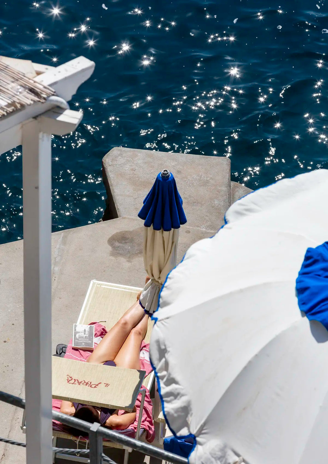 Person lying on a sunbed under a blue sky with a blue umbrella and white lounge chair and starry sunbursts in the water.