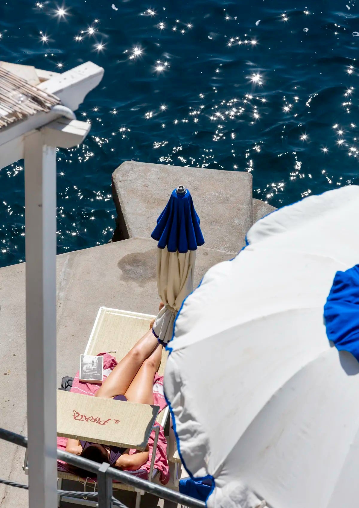 Person lying on a sunbed under a blue sky with a blue umbrella and white lounge chair and starry sunbursts in the water.