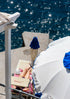 Person lying on a sunbed under a blue sky with a blue umbrella and white lounge chair and starry sunbursts in the water.