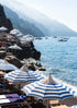 Seaside scene with blue and white striped umbrellas, rocky cliffs, and mountains in the background.