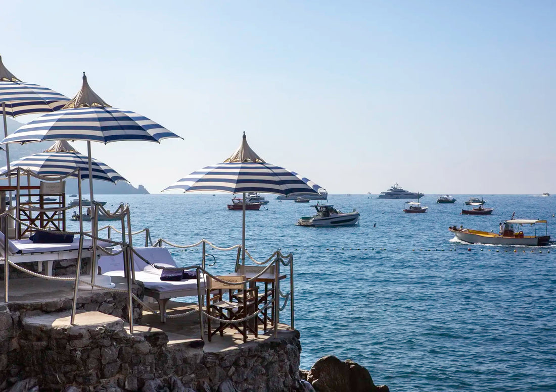 Beachside lounge area with striped umbrellas and chairs overlooking a calm sea with boats.