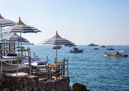 Beachside lounge area with striped umbrellas and chairs overlooking a calm sea with boats.