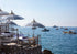 Beachside lounge area with striped umbrellas and chairs overlooking a calm sea with boats.