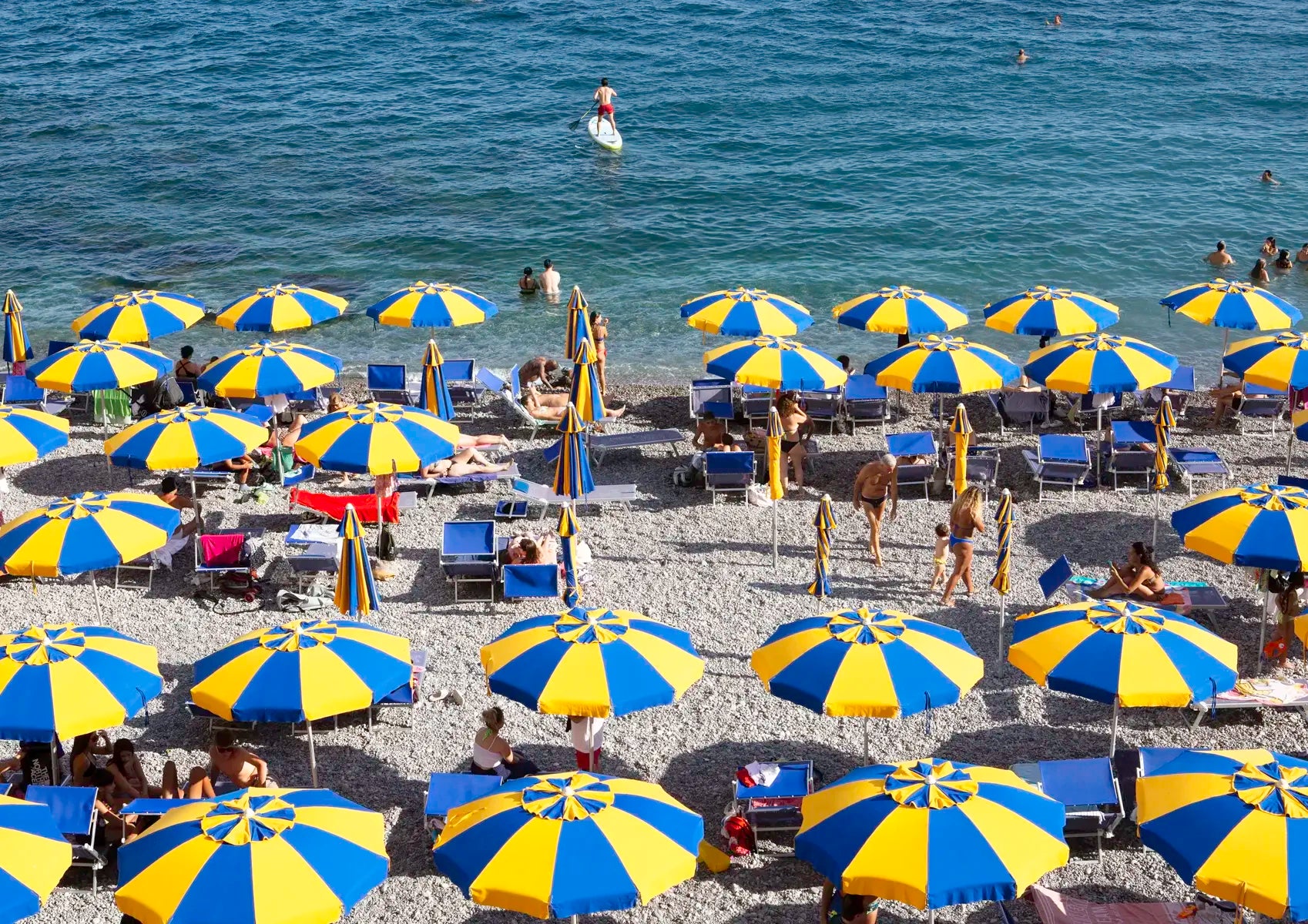Beach scene with numerous blue and yellow umbrellas on a pebble beach.