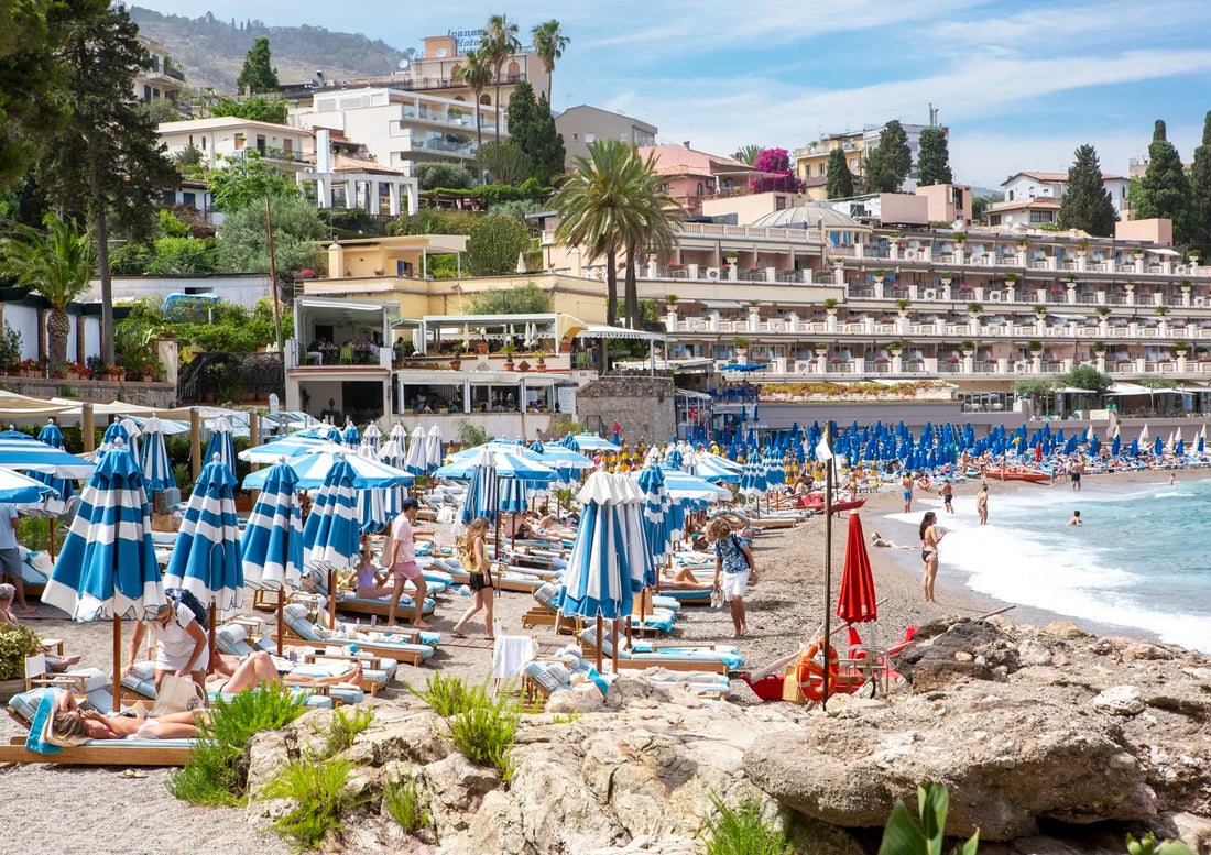 Beach scene with blue and white striped umbrellas, people, and buildings in the background.