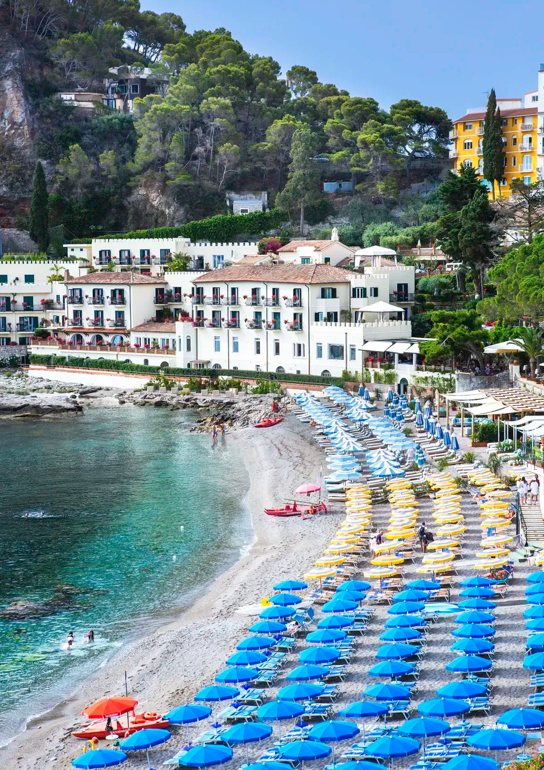 Beach scene with blue and yellow umbrellas, white buildings, and greenery.