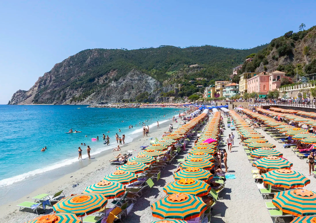 Beach scene with rows of colorful umbrellas and people on a sandy shore with mountains in the background.