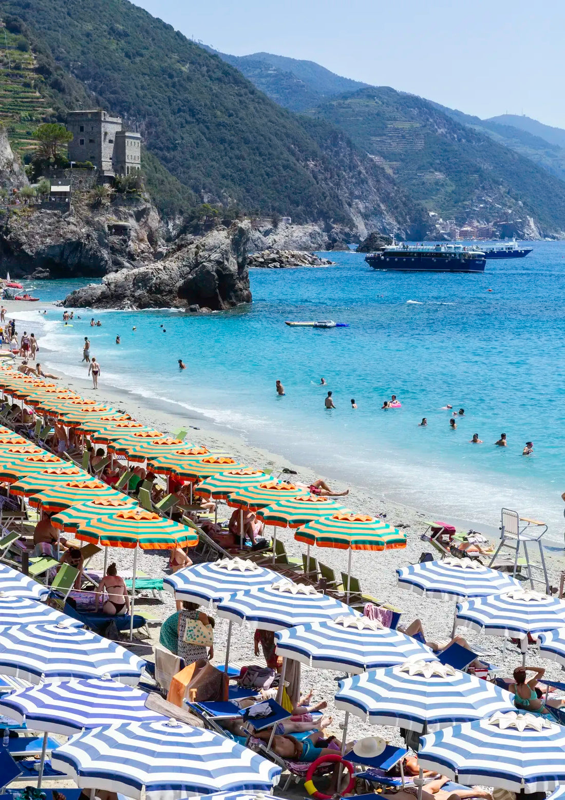 Beach scene with striped umbrellas, people, and boats near a mountainous coastline.