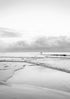Black and white beach scene with people walking on the horizon under a cloudy sky.