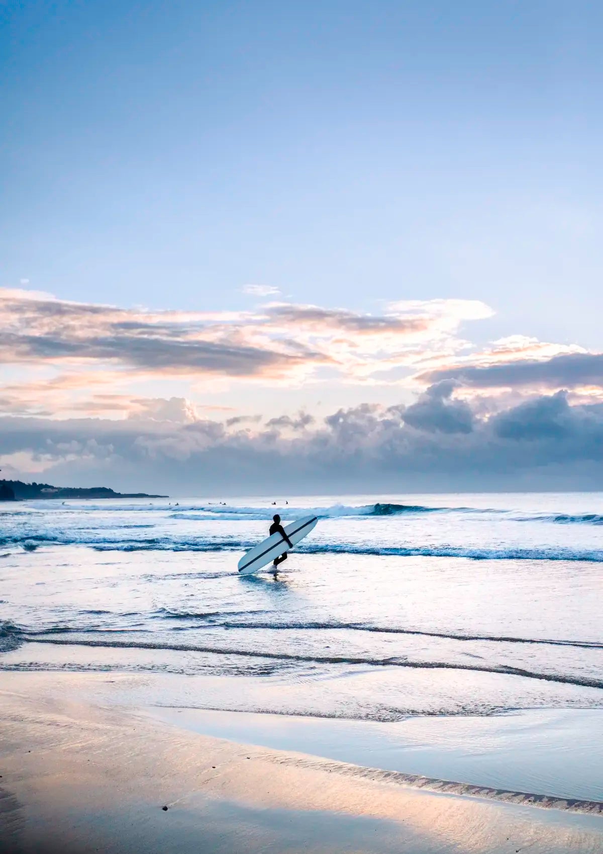 Person holding a surfboard walking along a beach with a cloudy sky.