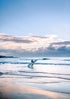 Person holding a surfboard walking along a beach with a cloudy sky.