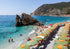 Beach scene with colorful umbrellas, people, and a rocky outcrop in the water.