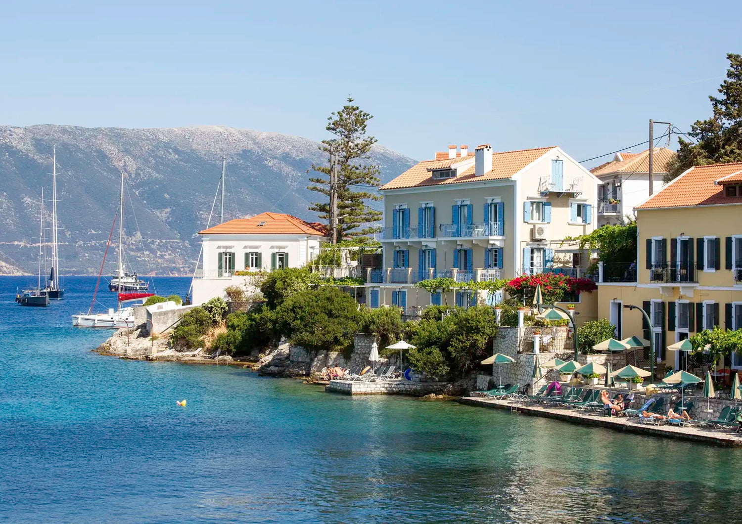Coastal scene with houses by the water, sailboats, and mountains in the background.