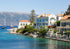 Coastal scene with houses by the water, sailboats, and mountains in the background.