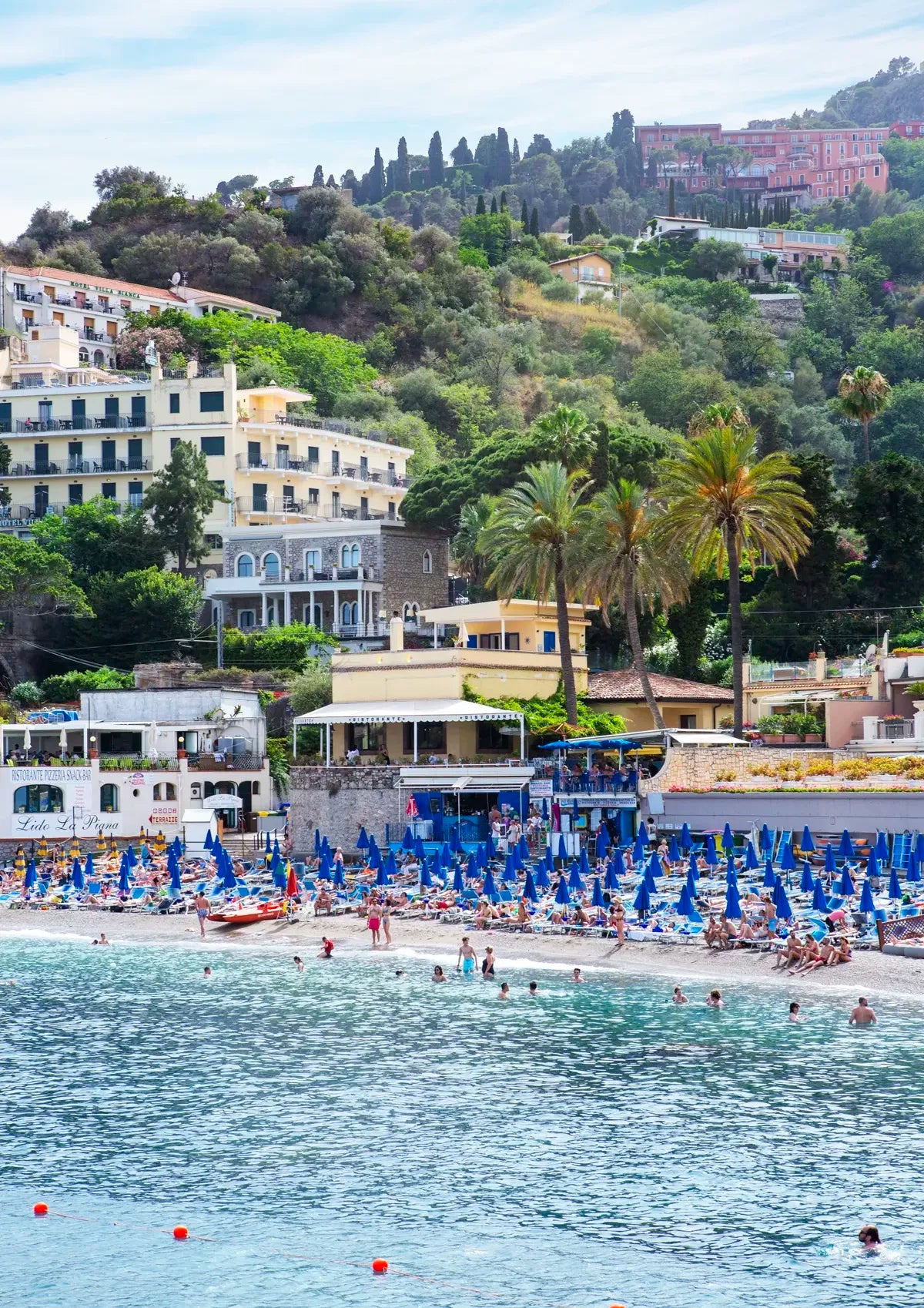 Beach scene with blue umbrellas, people, and buildings on a hillside.
